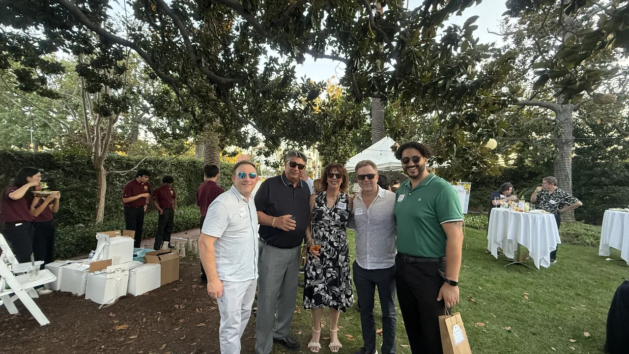 Several people stand together at an outdoor garden party, wearing sunglasses.