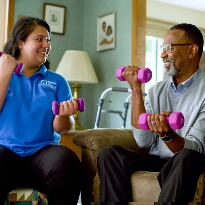 A female staff member in a blue uniform holds dumbbells with a male client during indoor physical therapy exercises.