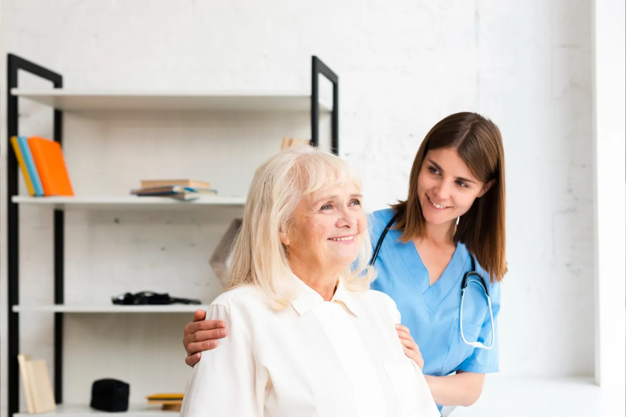 A caregiver in blue scrubs supports an elderly woman in a bright indoor space, facing her with a friendly expression.